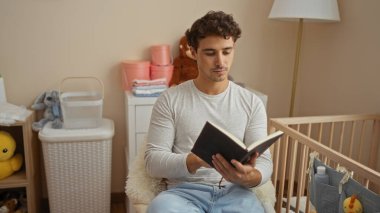 Genç adam, bebek odasında rahat bir bebek odası ve oyuncaklarıyla kitap okuyor, düşünceli bir ifade ve günlük giysiler sergiliyor, rahatlatıcı ve besleyici bir atmosfer yaratıyor..