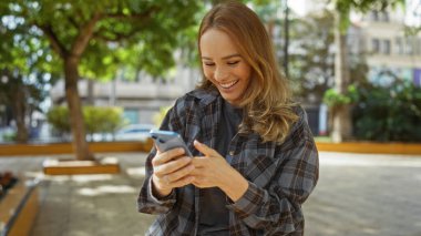 Şehir parkında akıllı telefon kullanırken gülümseyen bir kadın çevresini çevreleyen ağaçlarla güneşli bir günde mutluluğu ve rahatlığı yakalıyor..
