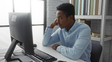 Genç adam, bilgisayar ve raflarla dolu bir ofis ortamında mavi gömlekli, profesyonel ve yoğun bir atmosferi olan işine odaklanmış..