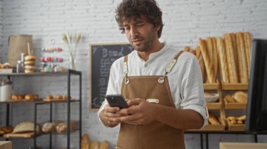 Genç bir adam fırında akıllı telefon kullanıyor. Etrafı taze ekmek ve pastalarla çevrili. Önlük ve beyaz gömlek giyiyor.