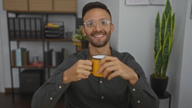 Sakallı genç bir İspanyol, sarı kupa tutuyor ve bir ofis ortamında gülümsüyor. Etrafı kitaplıklarla ve ev bitkileriyle çevrili..