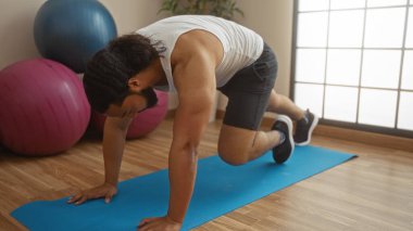 Mavi hasırın üzerinde egzersiz yapan bir adam, yakınlarda egzersiz topları, ahşap döşemeli ve doğal ışıklandırmalı bir sağlık merkezinde zindelik ve güç gösterisi yapıyor..