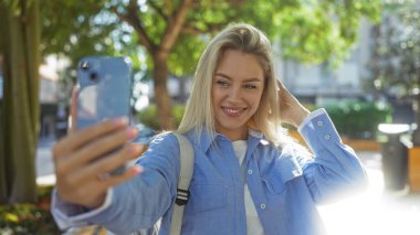 Güneşli bir günde şehir parkında selfie çeken bir kadın ağaçlarla ve şehir geçmişiyle sarışın güzelliğini sergiliyor, canlı bir açık hava yaşam tarzı ve gençlik ruhu yakalıyor..