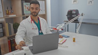 Veteriner kliniğinde çalışan genç İspanyol bir adam, steteskoplu beyaz önlük giyiyor, dizüstü bilgisayar kullanıyor, arkasında tıbbi ekipmanlar var..