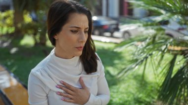 Göğsü göğsünde bir kadın güneşli bir parkta şehir yaşamını düşünüyor şehir yeşillikleriyle çevrili açık havada huzurlu bir anı yakalıyor..