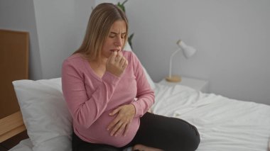 Huzurlu bir yatak odasındaki yatakta oturan hamile bir kadın, pembe bir bluz giyiyor, karnını nazikçe içeride tutuyor, evde bir bebek beklerken huzurlu bir anı yakalıyor..