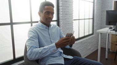 Kapalı bir odada oturan genç bir adam, elinde parlak bir ofis odasında odaklanmış bir ifadeyle uzun pencereler ve etrafında modern bir masa düzeniyle profesyonelliği aktarıyor..