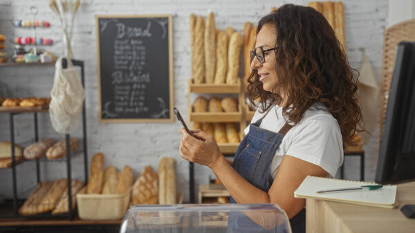 Woman in bakery wearing an apron uses smartphone surrounded by fresh bread and pastries, showcasing a cozy interior and her concentration on the business activities.