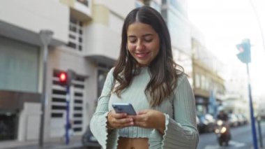 Kentsel caddede akıllı telefon kullanırken gülümseyen genç bir kadın modern bir bağlantı ve mutluluk hissi taşıyor..