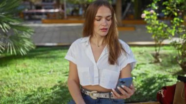 Şehir parkında çim üzerinde akıllı telefon kullanan, günlük beyaz gömlek giyen yeşil ve canlı arka planda güneşli bir günün tadını çıkaran bir kadın..