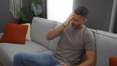 Kanepede oturan İspanyol bir adam modern bir oturma odasında yorgun görünüyor, evinde rahat ve içsel bir anı yakalıyor..