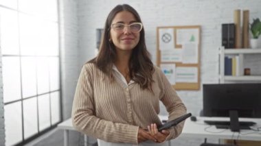 Parlak ofis ortamında elinde tabletle gülümseyen ve arka planda modern mobilya ve ilan panosu olan bir kadın, kapalı alanda profesyonel bir çalışma ortamı olduğunu gösteriyor..