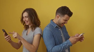 Çift sırt sırta vererek izole edilmiş sarı arka planda akıllı telefonlara bakıyor, modern teknoloji ve bağlantı sergiliyor.