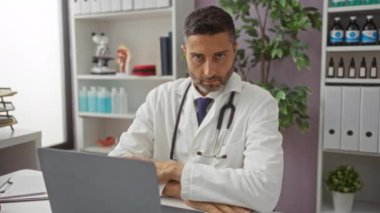 Steteskoplu beyaz önlüklü doktor klinikteki masada oturuyor ve arka planda rafları ve bitkileri olan bir tıbbi ortamda dizüstüne bakıyor.