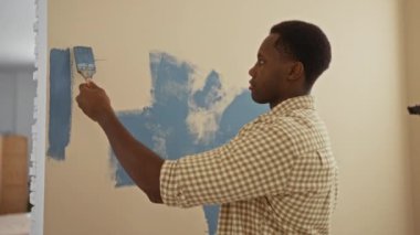 Oturma odasında duvar boyayan, kareli gömlek giyen, işine odaklanmış, modern bir ev iç atmosferi yaratan, ev geliştirme becerilerini gösteren genç bir adam..