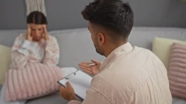 Bir adam, yastıklı ve hafif ışıklandırmalı rahat bir oturma odasında elinde bir pano tutarak, başı dertte olan bir kadını dinliyor.