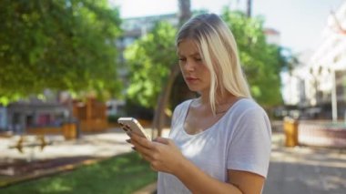 Güneşli bir günde şehir parkında akıllı telefon kullanan bir kadın ağaçlar ve binalarla çevrili asfalt bir yolda dikilirken endişeli bir yüz ifadesi sergiliyor.