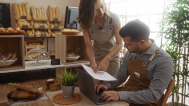Erkek ve kadın fırıncılar bir pastanenin içinde birlikte çalışıyorlar, arka planda raflar dolusu ekmekle birlikte ortak çalışma ortamını gösteren kağıtları tartışıyorlar.
