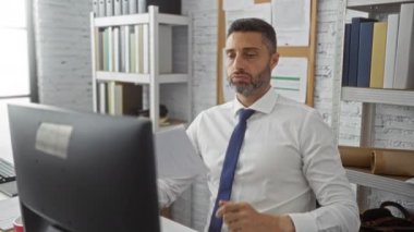 Ofiste çalışan, belgeleri tutarken kravat ayarlayan, kitap ve bilgisayarlarla modern çalışma alanında stresli görünen, profesyonel bir iç atmosfer yaratan bir adam..