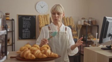 Beyaz bir bluz ve gri bir önlükle arka planda ekmek rafları olan sıcak bir fırında duran pastanede kruvasan düzenleyen bir kadın.