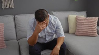 Oturma odasında gri bir kanepede oturan genç adam pembe yastıklı, stresli görünüyor..
