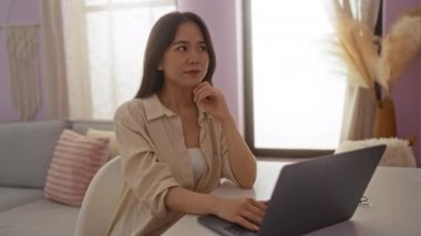 Asya 'lı bir kadın Çin' de evinde dizüstü bilgisayarla oturma odasında düşünceli ve yaratıcı bir anın altını çiziyor..