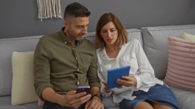 Rahat oturma odasında akıllı telefon ve tablet kullanan çift, modern aile ortamlarında sık sık görülen teknolojiden anlayan kapalı bir yaşam tarzını gösteriyor..