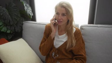 Telefondan konuşan bir kadın rahat bir dekorasyonla rahat ve konuşkan görünen rahat bir apartman dairesinde koltukta oturuyor.