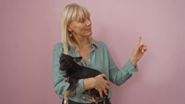 Orta yaşlı sarışın kadın ve tişört giyen sevimli köpek, ayakta ve mutlu bir şekilde yüzü soyutlanmış pembe arka planda gülümsüyor.