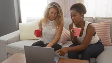 Kadınlar rahat bir oturma odasında kahve içerken konuşuyor, bir kanepede oturuyor, dizüstü bilgisayarla ahşap bir masada oturuyor.