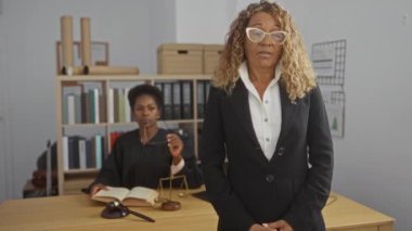 Siyah takım elbiseli kadın, hukuk kitapları ve terazilerle bir büro ortamında oturan bir yargıcın önünde duruyor.