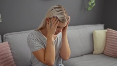 Oturma odasında oturan genç sarışın kadın, elleri şakaklarında stresli görünüyor.