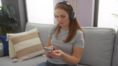 Kanepede dinlenen, kulaklık takan ve modern dekorlu rahat bir oturma odasında akıllı telefon kullanan bir kadın boş zaman aktiviteleri için huzurlu bir ev ortamı öneriyor..