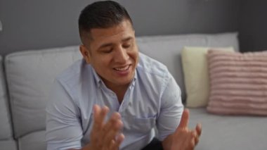 Arka planda dekoratif yastıklar ve gri bir kanepeyle oturma odasında konuşan genç bir adam.