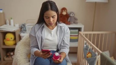 Akıllı telefon kullanan bir kadın rahat yatak odasında rahat oyuncaklar ve beşikle ürün etiketi okurken besleyici bir ortam öneriyor..