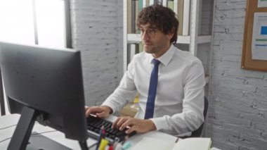 Beyaz gömlekli genç adam modern bir ofiste bilgisayar başında çalışıyor. Etrafı kitaplarla ve grafiklerle çevrili bir şekilde memnuniyet ve neşe ifade ediyor..