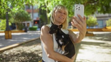 Güneşli bir günde şehir parkında köpeğiyle selfie çeken yeşil ağaçlı bir kadın.