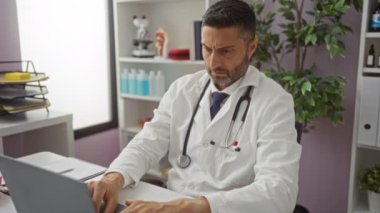 Ofiste steteskop takan bir doktor laptopta çalışıyor ve tıbbi malzemeler ve saksı bitkileriyle çevrili bir ifadeyle çalışıyor..