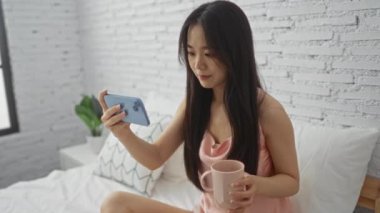 Kahve içen bir kadın, rahat bir yatak odasında beyaz tuğlalı duvarlara bakarak sakin bir sabah rutinini somutlaştırıyor.