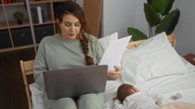 Dizüstü bilgisayarda çalışan bir kadın rahat yatak odasında uyuyan bir bebekle yatakta otururken, ev ortamında iş ve aile hayatının dengesini vurguluyor..