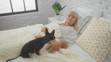 Beyaz dekorlu ve geometrik desenli yastıklı modern bir yatak odasında bir chihuahua köpeğiyle yatakta yatan olgun bir kadın.
