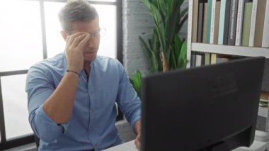Ofisteki genç adam gözlüklerini çıkartıyor ve elleri yüzü üzerinde dinleniyor. Arka planda kitap ve bitkilerin olduğu bir masada otururken yorgun ya da stresli görünüyor..