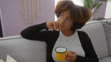 Modern oturma odasında, koltukta uzanan, sarı fincanı tutan kıvırcık saçlı ve gözlüklü bir kadın, evde tefekkür ve rahatlık sergiliyor..