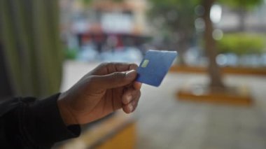 Şehir parkında, bulanık şehir geçmişine sahip mavi kredi kartı tutan adam açık hava finansal faaliyetlerini ve modern yaşam tarzını yansıtıyor..