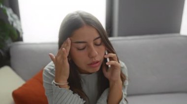 Telefonda konuşan kadın, evinde, oturma odasındaki koltukta oturmuş, eli alnında ve esmer saçlı stresli bir şekilde konuşuyor..