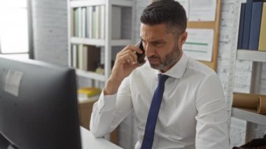Modern ofiste kitaplıklarla telefonla konuşan, iş iletişimine odaklanan, beyaz gömlek ve mavi kravat takan, profesyonel bir ortamda ciddi bir ifade sergileyen bir adam..