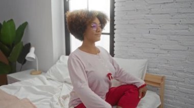 Kıvırcık saçlı bir kadın modern bir iç tasarımla rahat bir yatak odasında ayakta duruyor ve esniyor. Evde rahat bir sabah atmosferi sergiliyor..
