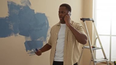 Yeni dairenin oturma odasında telefonla duvar boyayan bir adam arka planda merdiven kullanarak yenilemeyi öneriyor..