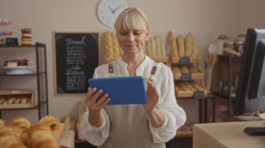 Fırında çeşitli ekmek somunları ve hamur işleriyle çevrili bir tablet kullanan bir kadın.