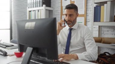 Modern bir ofiste çalışan bir adam, kitaplıklar ve evrak işleriyle çevrili bir bilgisayarda çalışırken telefonla konuşuyor, profesyonel bir iş ortamını vurguluyor..